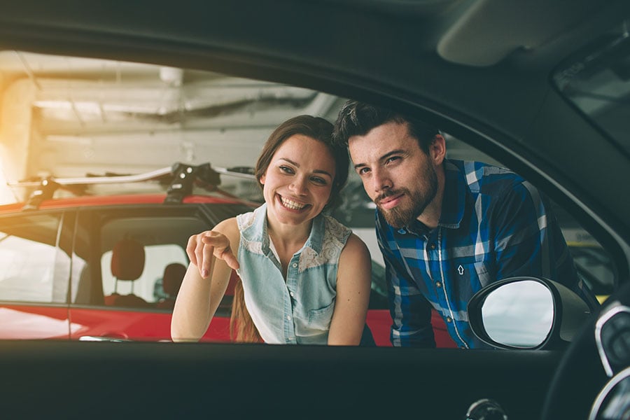a young couple wonders how to save money when buying a car as they look in the window of a vehicle they want to purchase