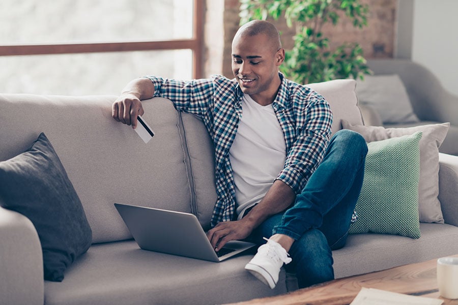A young man sits on a couch with his credit card and laptop mapping out his credit card payoff plan.